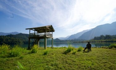 A serene scene with a man relaxing by a lake under a clear sky and observation tower.